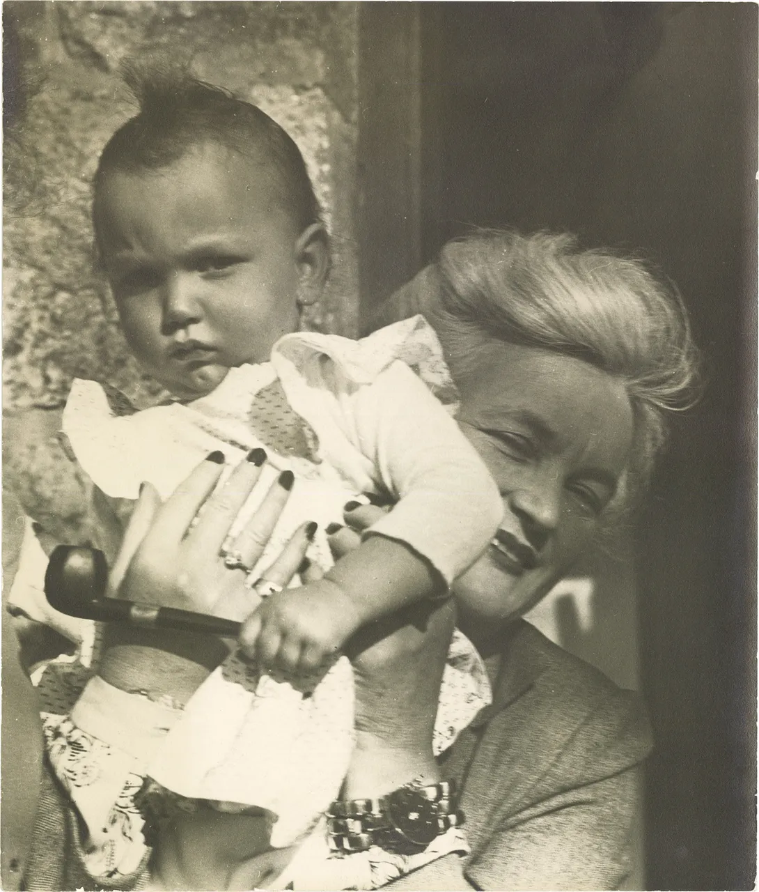 An older woman holding a small child who holds a tobacco pipe, outdoors.